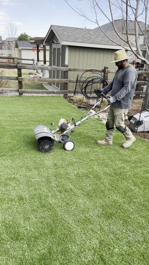 A crew member in a straw hat pushing a power broom across a newly installed artificial lawn, Mead–Firestone area, Colorado