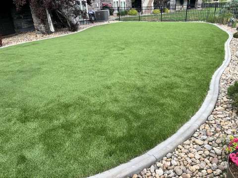 An artificial turf lawn with a curved concrete curb and river rock, a black metal fence behind