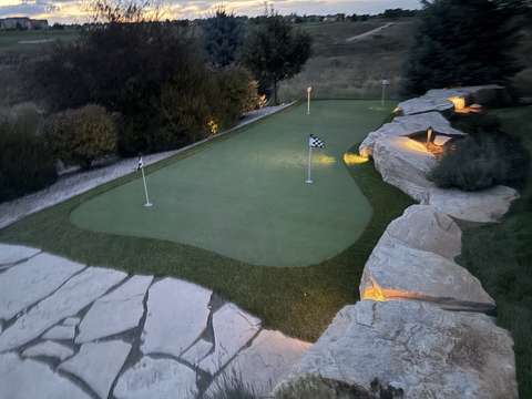 A putting green at dusk with checkered flags, boulders lit by landscape lights and a flagstone patio, near Windsor, Colorado