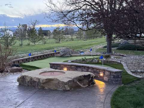 A backyard putting green with blue flags beside a stone fire pit and lit seat wall, the Front Range on the horizon, near Berthoud, Colorado
